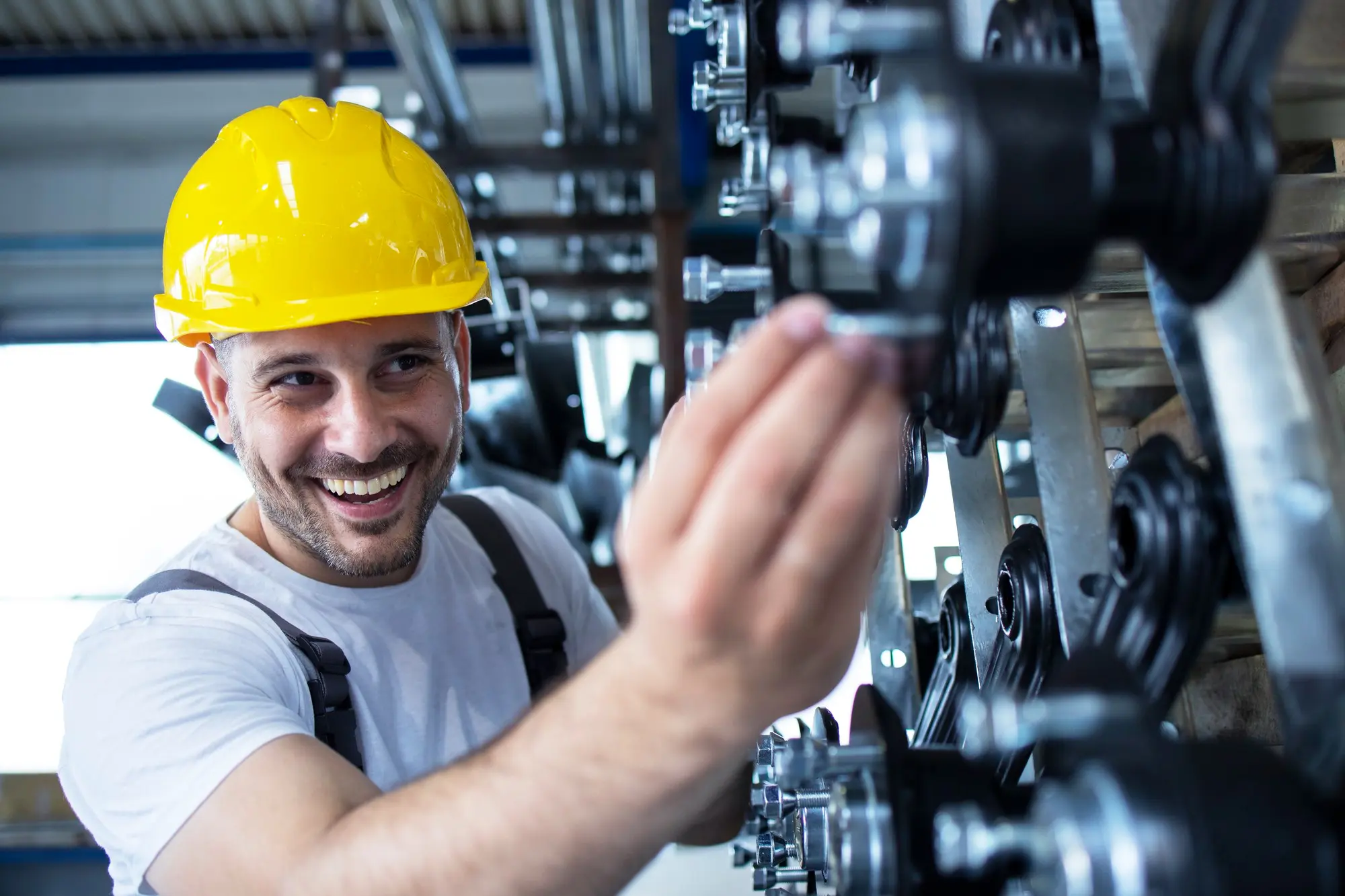 Trabalhador industrial, de camiseta branca e capacete amarelo, inspeciona equipamento em um sistema de produção, representando uma das atividades da engenharia de manutenção. Sorrindo, ele segura um componente cilíndrico enquanto o examina.